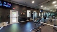 This fitness center offers a comprehensive workout environment designed for functionality and performance. The interior is defined by deep charcoal-painted walls trimmed in crisp white, creating a bold and motivating atmosphere. Durable rubber flooring with a speckled finish covers the entire space, providing both safety and resilience underfoot.

The facility is outfitted with a strong selection of professional-grade equipment, including a Bodycraft cable machine, a TRUE functional training rig, a full dumbbell rack ranging across multiple weight increments, and several adjustable workout benches. A wall-mounted flat-screen television keeps users informed and entertained during their sessions.

Full-length mirrors line an entire wall, visually expanding the space while allowing users to monitor their form. Recessed ceiling lights provide even, bright illumination throughout, complemented by generous natural light streaming in through multiple white-framed windows fitted with blinds. French-style glass doors along one wall offer additional outdoor views and easy access.

Crown molding adds a refined architectural touch, elevating the overall aesthetic beyond a typical gym setting. The layout is open and well-organized, allowing users to move freely between stations. This fitness center combines practical amenities with a polished design to deliver an effective and inviting exercise environment.