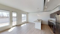 This single-floor, two-bedroom, one-bathroom residence offers a well-designed open-concept layout that maximizes both functionality and style. The main living and kitchen area is immediately welcoming, featuring light-toned hardwood-style flooring that extends throughout, creating a cohesive and airy feel. Large windows and a sliding glass door flood the space with natural light, offering views of the surrounding exterior and enhancing the overall sense of openness.

An exposed metal ductwork element runs along the ceiling, lending an industrial-chic character to the otherwise clean, minimalist aesthetic. Recessed lighting is complemented by stylish pendant lights suspended above a kitchen island, which features a granite-style countertop and an integrated sink. The crisp white cabinetry and bright walls contribute to the fresh, contemporary atmosphere.

The open layout seamlessly connects the kitchen and living areas, making the space feel expansive and versatile. The two bedrooms provide comfortable private retreats, while the single bathroom rounds out this thoughtfully designed unit, ideal for those seeking a modern, light-filled living environment.