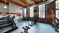 Located at 1531 S Eighth St in St. Louis, MO, this fitness center occupies a single floor and showcases a compelling blend of historic industrial architecture and modern exercise amenities. The space is defined by its striking exposed brick walls, warm wood-beam ceilings with visible timber framing, and large multi-pane windows that flood the room with natural light and offer views of the surrounding neighborhood. Distinctive concrete columns punctuate the interior, reinforcing the building's industrial character, while exposed ductwork running along the ceiling adds to the utilitarian aesthetic. A bold red accent stripe along the walls provides a dynamic visual contrast to the neutral tones throughout.

The exercise room is well-appointed with a comprehensive range of equipment, including multiple treadmills, elliptical machines, a cable strength-training system, free weights, and a weight bench. Durable textured rubber flooring covers the workout area, providing both safety and functionality. A large wall mirror expands the sense of space and supports strength-training activities. Pendant lighting fixtures hang from the ceiling, supplementing the ample natural light. A wall-mounted television is also present, enhancing the workout experience. The overall ambiance is energetic yet grounded, combining the character of a historic building with the practical demands of a modern fitness facility.