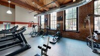 Located at 1531 S Eighth St in St. Louis, MO, this fitness center occupies a single floor and showcases a compelling blend of historic industrial architecture and modern exercise amenities. The space is defined by its striking exposed brick walls, warm wood-beam ceilings with visible timber framing, and large multi-pane windows that flood the room with natural light and offer views of the surrounding neighborhood. Distinctive concrete columns punctuate the interior, reinforcing the building's industrial character, while exposed ductwork running along the ceiling adds to the utilitarian aesthetic. A bold red accent stripe along the walls provides a dynamic visual contrast to the neutral tones throughout.

The exercise room is well-appointed with a comprehensive range of equipment, including multiple treadmills, elliptical machines, a cable strength-training system, free weights, and a weight bench. Durable textured rubber flooring covers the workout area, providing both safety and functionality. A large wall mirror expands the sense of space and supports strength-training activities. Pendant lighting fixtures hang from the ceiling, supplementing the ample natural light. A wall-mounted television is also present, enhancing the workout experience. The overall ambiance is energetic yet grounded, combining the character of a historic building with the practical demands of a modern fitness facility.