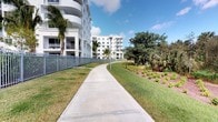 This outdoor space features a smooth, wide concrete walking path that curves gracefully through a meticulously maintained landscape. Flanking the pathway on one side is a well-manicured lawn bordered by a sleek metal fence that delineates the property boundary. On the opposite side, richly mulched garden beds are planted with a variety of low-growing tropical and native plants, including palms, ornamental grasses, and subtropical ground cover, creating vibrant layers of texture and color. Mature oak and palm trees provide natural shade and visual depth along the path's edge, adding to the lush, tropical character of the setting. Pedestrian-scale light poles are positioned at intervals along the walkway, ensuring safe and pleasant use during evening hours. The backdrop features a modern, multi-story building with a clean white exterior, curved balconies, and large windows, reflecting a contemporary architectural aesthetic consistent with Florida's coastal design sensibility. The well-kept grounds, abundant greenery, and open sky create an inviting, park-like atmosphere that encourages outdoor activity and leisurely strolls within the community.