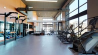 This contemporary fitness center is designed to deliver a premium workout experience within a thoughtfully organized, open-plan layout. The space is anchored by dramatic floor-to-ceiling windows along multiple walls, flooding the interior with abundant natural light and offering views of the surrounding urban neighborhood. Soaring ceilings with exposed ductwork give the facility an industrial-modern aesthetic, complemented by warm terracotta-toned accent walls and durable rubberized flooring throughout.

The cardio zone is generously appointed with a full row of Matrix treadmills, elliptical machines, and stationary bikes, all positioned to take advantage of the outdoor views. A dedicated strength training area features a comprehensive cable and multi-station machine setup, with large wall-mounted mirrors enhancing the sense of space and supporting proper form. Rowing machines provide additional cross-training options in an open floor zone.

A functional training section is equipped with a rig system supporting pull-up bars, TRX suspension trainers, and an organized rack of kettlebells and medicine balls, catering to dynamic and bodyweight-based workouts. A separate glass-enclosed studio area offers space for cycling classes or focused group training sessions. Recessed and linear overhead lighting complements the natural light, ensuring the facility remains well-lit at all hours. The overall design is clean, spacious, and purpose-built for a diverse range of fitness activities.