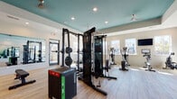 Located at 1079 Beckstead Ct in Denver, NC, this fitness center is a thoughtfully designed, single-floor facility that offers a comprehensive workout experience. The space is immediately distinguished by its striking teal accent ceiling, which adds a vibrant pop of color against the neutral white walls and warm hardwood-style flooring throughout.

Natural light pours in through multiple large windows, creating an open and energizing atmosphere. The cardio section is well-appointed with a row of treadmills, ellipticals, and a stationary bike, complemented by a wall-mounted television for entertainment during workouts. A rowing machine adds further variety to the cardio offerings.

The strength training area is equally robust, featuring a multi-functional cable machine, a squat rack with weight plates, a flat bench, a power tower for bodyweight exercises, and plyo boxes for functional training. Free weights and additional accessories are neatly organized on shelving units.

A large floor-to-ceiling mirror spans one wall, enhancing the sense of space and allowing users to monitor their form. The mirror reflects the full depth of the facility, including motivational fitness-themed text displayed on the teal accent wall. Recessed lighting and ceiling fans provide consistent, comfortable illumination throughout. The space is accessible via a glass-panel entry door, making for a clean and welcoming entrance.