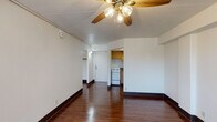 This single-floor unit is defined by its clean, straightforward layout and practical design. The main living area is the focal point of the space, showcasing rich, dark hardwood floors that extend throughout. A wide horizontal window runs along one wall, allowing natural light to fill the room while offering views of the surrounding exterior. A ceiling fan provides air circulation, and a wall-mounted PTAC unit ensures climate control. Track lighting along the ceiling adds a functional lighting option to the open space.

The kitchen is tucked adjacent to the main living area and is outfitted with warm wood cabinetry extending to the ceiling, providing generous storage. Dark laminate countertops offer ample workspace, and the kitchen includes a white electric range, a refrigerator, and a stainless steel sink set into an L-shaped counter configuration. Hardwood flooring continues into this area, maintaining visual continuity.

The bathroom is finished in a clean, neutral palette with white tile surrounding a full bathtub and shower combination. A wood-toned vanity with an integrated sink, a wall-mounted mirror with overhead lighting, and mosaic tile flooring complete the space. The unit also features a closet area near the entryway, adding practical storage.