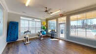 Located at 321 S Swing Rd in Greensboro, NC, this single-story fitness center at Comet Greensboro is thoughtfully divided into distinct workout zones, each designed to support a comprehensive exercise experience. Crown molding, neutral gray walls, and ceiling fans lend a polished, cohesive aesthetic throughout the facility.

The cardio zone features multiple TRUE-brand treadmills, an elliptical, and a rowing machine arranged along windowed walls that allow ample natural light to fill the space. A wall-mounted flat-screen television provides entertainment during workouts. Durable rubber flooring in this area ensures safety and comfort underfoot.

The strength training zone is equipped with a power rack, adjustable benches, and a full dumbbell rack displayed in front of floor-to-ceiling mirrors, creating a spacious, well-lit environment ideal for resistance training. Additional overhead lighting and windows further brighten the room.

A separate studio zone features rich hardwood-style flooring and houses spinning bikes, stability balls in multiple sizes, exercise mats, and resistance accessories neatly organized along the walls. Glass-panel doors in this area open directly to an outdoor space, enhancing ventilation and providing views of the surrounding community. Together, these zones create a well-rounded, fully equipped fitness facility.