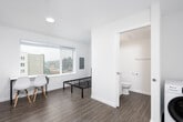 This compact studio unit offers a thoughtfully designed open-concept layout that efficiently combines living, sleeping, and dining areas within a single bright space. The main room features warm wood-tone flooring, crisp white walls, and a large window that fills the space with natural light while framing views of the surrounding urban streetscape.

The kitchen is a standout feature, outfitted with a full suite of stainless steel appliances, including a full-size refrigerator, electric range, over-the-range microwave, and dishwasher. White flat-front cabinetry extends to the ceiling, maximizing storage, while the clean white countertops and single-basin sink complete a sleek, modern aesthetic.

A dedicated dining area with a compact table and chairs sits adjacent to the kitchen, making efficient use of the available space. The entry area accommodates an in-unit washer, a practical convenience that adds significant value to the unit.

The bathroom is finished with wood-look flooring, a white vanity, toilet, and a shower-tub combination, maintaining the clean and minimal design found throughout the rest of the unit. Overall, the space presents a cohesive, modern aesthetic well-suited for efficient urban living.