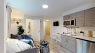 This thoughtfully designed studio unit maximizes every inch of its single-floor layout, offering a bright and functional living environment with a clean, contemporary aesthetic throughout. Light wood-look flooring extends seamlessly across the entire space, tying the open-plan interior together with a warm, cohesive feel.

The main living area serves a dual purpose as both a sleeping and lounge space, anchored by a daybed-style sofa positioned beneath a large window that invites natural light. A built-in open closet provides ample hanging and shelving storage, while an additional wall-mounted television adds convenience.

The kitchenette is efficiently arranged with modern, wood-grain laminate cabinetry, a white countertop, a stainless steel sink, an induction cooktop, a microwave, and a compact refrigerator. A wall-mounted utensil rail keeps essentials close at hand. A small dining nook with a fold-out table and chair sits adjacent to the kitchen, making the most of the available footprint.

The private bathroom features light-toned walls, wood-look flooring, a wall-mounted sink with a framed mirror, a toilet, and a small frosted window for natural ventilation. The unit's entry foyer provides a clear transition into the living space, complete with a coat hook rack near the front door. Recessed ceiling lighting throughout ensures a clean, modern finish.