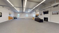 This single-floor multipurpose space combines industrial architectural character with functional amenities suited for events, meetings, and gatherings. The centerpiece is a generously proportioned open hall defined by soaring ceilings with exposed steel trusses running its length, illuminated by rows of linear fluorescent lighting. Speckled carpet flooring extends throughout, and crisp white walls provide a clean, neutral backdrop. Multiple windows allow natural light to filter in, adding brightness to the expansive interior.

A lounge area at one end of the main hall features a gas fireplace with a white mantle, a wall-mounted flat-screen television, leather seating, and a collection of framed artwork. Folding chairs and a wooden storage cabinet are available for flexible seating arrangements. A dedicated conference room accommodates a full boardroom-style table surrounded by ergonomic chairs, with natural light from a large window and wall-mounted décor providing a polished, professional atmosphere.

A step-down kitchen area is equipped with wooden cabinetry, a black refrigerator, stove, microwave, and sink, supporting catering and event needs. The overall ambiance is clean, well-lit, and adaptable, making the space suitable for a wide range of uses.