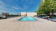 Located at 111 Towne St in Stamford, Connecticut, this expansive outdoor amenity deck offers a thoughtfully designed environment for relaxation and recreation. At its centerpiece sits a large rectangular swimming pool with stainless steel entry rails, surrounded by an extensive sun deck lined with rows of lounge chairs and umbrellas — ideal for taking in the open sky and fresh air. The pool area is enclosed by sleek black iron fencing and accented by mature trees that provide natural shade along the perimeter. Lush, professionally maintained garden beds featuring hydrangeas, ornamental grasses, and seasonal plantings are interspersed throughout the terrace, adding vibrant color and a resort-like ambiance. A dedicated lounge zone showcases a fire pit surrounded by Adirondack chairs in a bold aqua hue, creating an inviting social gathering space. Nearby, outdoor grilling stations offer a functional complement to alfresco entertaining. A hammock station provides a peaceful retreat, while wide-format stone pavers unify the entire deck with a clean, contemporary aesthetic. The terrace also offers open views of the surrounding Stamford skyline. A curved, glass-facade building structure anchors the space architecturally, connecting the amenity areas with a sense of cohesion and modern design.