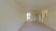 This single-floor space presents an open and airy layout finished in soft, neutral tones throughout. The main living area is generously proportioned, featuring light beige carpeting that extends across the floor, contributing to a clean and understated aesthetic. Crown molding runs along the upper perimeter of the walls, adding a subtle architectural detail that enhances the overall refinement of the space.

A central pendant light fixture hangs from the ceiling, providing a focal point in the room while offering functional overhead lighting. Natural light enters through a multi-pane window, brightening the interior and creating a welcoming atmosphere. The walls are painted in a warm off-white tone, complementing the light carpeting and contributing to the sense of openness.

Toward the back of the space, two white interior doors are visible, leading to additional rooms or closet areas. An electrical panel is mounted on the wall nearby, and a staircase railing is partially visible to the right, suggesting access to additional areas of the building. Overall, the space offers a clean, move-in ready environment with a simple and functional design.