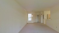 This single-floor space presents an open and airy layout finished in soft, neutral tones throughout. The main living area is generously proportioned, featuring light beige carpeting that extends across the floor, contributing to a clean and understated aesthetic. Crown molding runs along the upper perimeter of the walls, adding a subtle architectural detail that enhances the overall refinement of the space.

A central pendant light fixture hangs from the ceiling, providing a focal point in the room while offering functional overhead lighting. Natural light enters through a multi-pane window, brightening the interior and creating a welcoming atmosphere. The walls are painted in a warm off-white tone, complementing the light carpeting and contributing to the sense of openness.

Toward the back of the space, two white interior doors are visible, leading to additional rooms or closet areas. An electrical panel is mounted on the wall nearby, and a staircase railing is partially visible to the right, suggesting access to additional areas of the building. Overall, the space offers a clean, move-in ready environment with a simple and functional design.