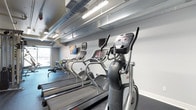 This fitness center offers a focused and functional workout environment designed to meet a variety of cardio training needs. The space features a lineup of modern cardio equipment, including multiple treadmills and elliptical machines, each outfitted with digital display consoles for tracking workout metrics. A rowing machine is also available, rounding out the cardio offerings for a comprehensive exercise experience. The interior design takes on an industrial aesthetic, with exposed silver ductwork running along the ceiling alongside fluorescent strip lighting that ensures the space is bright and well-lit throughout. The walls are finished in a clean, neutral white, contributing to an open and airy atmosphere. Dark rubberized flooring provides a practical, durable surface suited for high-traffic fitness use. A wall-mounted flat-screen television is positioned for easy viewing during workouts, adding an entertainment element to the exercise experience. A window along one wall allows for natural light to filter into the space, enhancing the overall ambiance. The single-floor layout is efficiently organized, making the most of the available footprint while maintaining clear, unobstructed pathways between equipment.