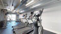 This fitness center offers a focused and functional workout environment designed to meet a variety of cardio training needs. The space features a lineup of modern cardio equipment, including multiple treadmills and elliptical machines, each outfitted with digital display consoles for tracking workout metrics. A rowing machine is also available, rounding out the cardio offerings for a comprehensive exercise experience. The interior design takes on an industrial aesthetic, with exposed silver ductwork running along the ceiling alongside fluorescent strip lighting that ensures the space is bright and well-lit throughout. The walls are finished in a clean, neutral white, contributing to an open and airy atmosphere. Dark rubberized flooring provides a practical, durable surface suited for high-traffic fitness use. A wall-mounted flat-screen television is positioned for easy viewing during workouts, adding an entertainment element to the exercise experience. A window along one wall allows for natural light to filter into the space, enhancing the overall ambiance. The single-floor layout is efficiently organized, making the most of the available footprint while maintaining clear, unobstructed pathways between equipment.