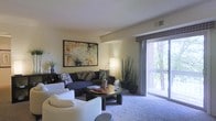 This single-floor residence offers a thoughtfully arranged living space that balances comfort and style. The living room serves as the centerpiece of the home, featuring plush wall-to-wall carpeting that adds warmth underfoot. A large sliding glass door floods the room with natural light while framing views of the surrounding greenery and providing direct access to an outdoor balcony — a standout feature that connects the interior to the natural environment outside.

The living area is generously proportioned, comfortably accommodating a sectional sofa, accent armchairs, and a central coffee table, creating an inviting layout ideal for both relaxing and entertaining. The neutral, light-toned walls provide a versatile backdrop, complemented by ceiling-height architectural details that lend subtle definition to the space. A bold abstract artwork anchors one wall, adding a creative focal point to the room.

Tasteful decorative touches, including indoor plants and ambient floor lighting, contribute to a calm and welcoming atmosphere. The room's design balances soft textures with clean lines, resulting in a space that feels both livable and curated. Overall, this residence offers a serene and comfortable living environment with a strong connection to the outdoors.