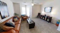 This single-floor space offers a comfortable and functional layout designed for everyday living. The living area serves as the centerpiece of the home, featuring soft wall-to-wall carpeting that adds warmth underfoot. Neutral-toned walls create a clean, versatile backdrop, while large windows along one wall invite ample natural light and provide a connection to the outdoors. A ceiling fan with integrated lighting is centrally positioned overhead, ensuring both comfort and illumination throughout the space. The room comfortably accommodates a generous seating arrangement, including a rich cognac leather sofa and matching armchair accented with colorful throw pillows, all oriented around a dark wood coffee table. Opposite the seating area, a media console provides practical storage and display space alongside a wall-mounted television. A dedicated desk and chair in the corner offer a functional workspace within the living area. Decorative touches, including a potted palm and table lamps, contribute to the inviting and lived-in ambiance of the space. The overall design balances practicality with comfort, making it a welcoming environment suitable for both relaxing and entertaining.