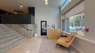 The lobby of East Eleven, located at 433 SE 13th Ave in Portland's vibrant SE neighborhood, presents a thoughtfully designed entrance that balances modern aesthetics with functional convenience. Upon entry, visitors are greeted by large floor-to-ceiling windows that flood the space with natural light and offer views of the surrounding tree-lined streetscape. The flooring throughout is finished in polished stone tiles, lending the space a refined, contemporary character.

A natural wood reception desk anchors the main entry area, accompanied by a comfortable mustard-yellow lounge chair and a minimalist side table, creating a welcoming seating nook. The soaring ceiling height adds a sense of openness and grandeur to the space. Substantial travertine-clad stairs with a sleek metal handrail lead to an upper level, beneath a striking multi-stem chandelier that serves as a dramatic focal point.

Adjacent to the staircase, a stainless steel elevator with lush tropical plant accents provides convenient vertical access. The mailroom features dark, geometric-patterned accent walls in deep teal, individual mailboxes, and automated package lockers beneath a bold cluster chandelier. Building signage notes amenities including a fitness center, rooftop, bike storage, and pet wash station.