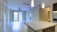 This single-floor space showcases an open-concept layout that seamlessly connects the kitchen and living areas. The kitchen features a granite-topped breakfast bar illuminated by sleek pendant lights, offering both a functional workspace and a casual dining spot. Track lighting overhead provides additional illumination throughout the kitchen zone.

The living area is finished with large-format light tile flooring that extends throughout the space, enhancing the sense of openness and reflecting natural light effectively. A ceiling fan is centrally positioned in the living area, ensuring comfortable airflow. Neutral-toned walls contribute to the clean, modern aesthetic throughout.

Floor-to-ceiling sliding glass doors lead to a private balcony, allowing natural light to fill the interior and providing outdoor views of the surrounding building and greenery. Vertical blinds offer adjustable light control and privacy.

A doorway visible from the main living area leads to additional rooms within the unit. The overall ambiance is bright, airy, and well-designed, with a cohesive contemporary style maintained through consistent finishes and thoughtful lighting choices.