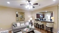 This single-story residence is designed with an open-concept layout that seamlessly connects the living area to the kitchen, creating a cohesive and functional space. The living room is warm and inviting, featuring neutral beige walls accented by elegant crown molding that adds a refined architectural touch. A five-blade ceiling fan with integrated lighting serves as a functional centerpiece overhead, ensuring comfort and illumination. A large decorative mirror with a geometric frame adorns one wall, enhancing the sense of depth in the room. The space accommodates a plush gray sofa dressed with patterned throw pillows, complemented by a rustic wooden coffee table that anchors the seating area.

An arched passthrough visually connects the living room to the kitchen, where rich, dark wood cabinetry pairs with dark stone countertops for a bold, classic look. A raised breakfast bar with bar-height seating provides a casual dining option. Pendant lighting above the kitchen area adds warmth and style. The dark hardwood flooring flows throughout, tying the spaces together while providing a grounding contrast to the lighter wall tones. The overall ambiance is cozy, polished, and well-appointed.