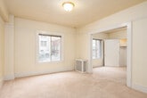 This single-floor unit offers a straightforward and functional layout designed to maximize livable space. The main living area is generously sized, featuring soft wall-to-wall carpeting, warm cream-toned walls, and classic flush ceiling lighting. Multiple windows throughout the space flood the rooms with natural light and provide views of the surrounding urban rooftop landscape and neighboring buildings, giving the space a connected city feel.

The layout includes what appears to be a primary room and an additional smaller room, each carpeted and outfitted with windows that offer natural light and outdoor views. The primary room includes a built-in closet with shelving, providing practical storage. A glass-panel interior door adds a subtle architectural detail, allowing light to pass between rooms.

The kitchen is compact and efficiently arranged, featuring white cabinetry both above and below laminate countertops, a stainless steel single-basin sink positioned beneath a window, and tile flooring. The space is well-suited for everyday meal preparation.

The bathroom is cleanly appointed with white subway tile surrounding a full bathtub and shower combination, a toilet, overhead cabinetry for storage, and a wall-mounted mirror cabinet. Tile flooring completes the practical, no-frills design.

Throughout the unit, white trim and molding details add a classic touch, while the consistent warm wall tones create a cohesive and neutral atmosphere.