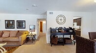This single-floor residence offers a thoughtfully arranged layout with distinct living, dining, kitchen, and sleeping areas, all unified by a warm and eclectic design aesthetic. The living room serves as a focal point, showcasing a striking exposed brick accent wall that adds texture and character to the space. Vaulted ceilings enhance the sense of openness, while large windows dressed in rich, copper-toned drape panels allow natural light to fill the room. Plush carpeting runs throughout the primary living and sleeping areas, contributing to the space's cozy ambiance.

The dining area features a large window with blinds and matching drapery, providing a bright setting for gatherings around a dark wood table and woven rattan chairs. Adjacent to the dining space, the galley-style kitchen is equipped with dark wood cabinetry, light stone-patterned countertops, a double basin sink, dishwasher, and under-cabinet appliances, all complemented by hardwood-style flooring.

The bedroom is comfortably sized, featuring a large window with draped curtains, soft carpeting, and refined decorative accents including framed artwork and ornamental wall pieces. A decorative black sideboard with woven baskets and a large ornate clock accentuates the transitional area between rooms, reinforcing the space's cohesive, classic-meets-eclectic character throughout.
