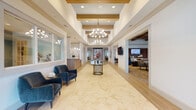 Located at 3650 RCA Blvd in Palm Beach Gardens, Florida, this single-story community space presents a sophisticated and welcoming environment defined by thoughtful architectural details and high-quality finishes. The grand entrance lobby sets the tone immediately, featuring expansive marble-look porcelain tile flooring, soaring tray ceilings accented with warm wood inlays, and an elegant chandelier that anchors the central corridor. A reception desk with natural wood paneling provides a polished welcome point, while a glass-top accent table with a decorative base adds a refined focal point to the main hall.

Adjacent to the lobby, a spacious lounge area showcases a striking coffered ceiling with contrasting wood beams painted in deep tones against a soft blue backdrop. This gathering space is furnished with versatile seating arrangements and opens to a secondary lounge featuring a stacked-stone fireplace with a wall-mounted television above, curated artwork, and a grand piano—creating a cultural and social hub within the space.

Natural light filters through large windows fitted with white window treatments throughout, contributing to the bright, airy ambiance. Additional areas include a quiet workstation nook and transitional corridors adorned with botanical-inspired artwork. The overall design balances coastal Florida warmth with contemporary elegance, creating a refined and functional communal environment.