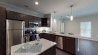 This well-appointed single-floor residence combines modern design with functional living. The centerpiece is a gourmet kitchen featuring dark espresso cabinetry, speckled granite countertops, a white subway tile backsplash, and a full suite of stainless steel appliances including a refrigerator, range, microwave, and dishwasher. Two kitchen islands provide generous prep and dining space, while elegant pendant lighting adds a contemporary touch. Crown molding accents the kitchen and living areas, adding a refined architectural detail throughout.

Rich, dark hardwood-style flooring flows seamlessly through the entire space, creating a cohesive and sophisticated aesthetic. The open living area is bright and spacious, with natural light streaming through blinds-fitted windows. French doors in the main living area open to a private balcony, enhancing the connection to the outdoors. Ceiling fans are featured in both the living room and bedroom, promoting comfort year-round.

The bedroom includes a walk-in closet with built-in shelving and racking, offering excellent storage. An additional room provides flexible use as a second bedroom or office. The bathroom is finished with granite countertops, dark cabinetry, and a full tub-shower combination. A laundry closet with louvered doors adds everyday convenience to this thoughtfully designed residence.