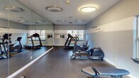Located at 4600 Big Tree Way in Greensboro, North Carolina, this well-appointed fitness center offers a comprehensive workout environment designed to meet a variety of exercise needs. The single-floor space is thoughtfully laid out, providing ample room to move freely between equipment without feeling cramped.

The facility is equipped with a strong selection of cardio machines, including treadmills, ellipticals, and a stationary bike, as well as a rowing machine for full-body conditioning. A multi-functional cable and weight machine anchors the strength-training area, complemented by a flat workout bench for additional exercises. Wall-mounted flat-screen televisions keep users entertained during their workouts.

A full wall of floor-to-ceiling mirrors lines one side of the room, enhancing the sense of space while allowing users to monitor their form. The fitness center features durable dark flooring suited for high-traffic activity, and the walls are painted in a calming two-tone gray palette accented by motivational typography reading "Everyday Is a Good Day to Work Out." Multiple windows along the perimeter flood the space with natural light and offer views of the surrounding wooded landscape. Overhead flush-mount lighting ensures the room remains bright at all times. A posted rules board and sanitation station underscore a commitment to cleanliness and community standards.