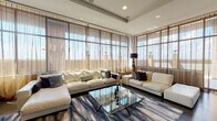 This refined single-floor space showcases a seamless blend of contemporary design and upscale finishes throughout. The expansive living area is the centerpiece, anchored by floor-to-ceiling windows wrapped in sheer, neutral-toned drapery that filters generous natural light while framing sweeping city views. Dark hardwood flooring runs throughout, complemented by a bold, abstract area rug that adds texture and color contrast beneath a sculptural glass-top coffee table. Plush white leather sectional sofas provide ample seating, while a striking driftwood-style floor lamp adds an organic accent to the refined setting. A recessed tray ceiling with integrated lighting further elevates the living space. Adjacent to the living area, a sleek fireplace set within a dramatic marble slab panel flanked by dark wood wall tiles serves as a sophisticated focal point. A classic leather lounge chair nearby adds a timeless design touch. The galley-style kitchen is efficiently appointed with warm wood cabinetry, white countertops, stainless steel appliances including a gas range and refrigerator, and an undermount sink — offering a clean, functional workspace. The overall ambiance is modern and polished, combining luxury materials with thoughtful spatial design.