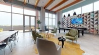 Located at 8446 S Dixie Hwy in Miami, FL, this single-floor amenity space is a contemporary and thoughtfully designed communal lounge. The interior is defined by soaring ceilings adorned with warm wood beam accents and recessed lighting, creating an inviting yet sophisticated atmosphere throughout.

Floor-to-ceiling windows line the perimeter, flooding the space with abundant natural light and offering sweeping views of the surrounding Miami skyline. The expansive main lounge area features multiple distinct seating arrangements, including clusters of sage green and neutral-toned lounge chairs, a cognac leather sofa, and a teal accent chair, all positioned around low tables atop textured rugs. A geometric, colorful accent wall provides a vibrant focal point alongside a mounted flat-screen TV and a game table. Open shelving units with brass poles serve as elegant dividers between zones.

The kitchen area is sleekly appointed with matte gray cabinetry, a large central island with a white marble-style countertop, vivid yellow bar stools, and eye-catching geometric patterned tile flooring in teal and yellow. High-end appliances, including a built-in oven and full-size refrigerator, complement the space. Polished concrete-style tile flooring flows throughout the main lounge, tying together this modern, light-filled, and versatile communal environment.