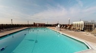 Located at 4400 East-West Hwy in Bethesda, Maryland, this rooftop pool facility offers a well-appointed aquatic amenity with sweeping open-sky views of the surrounding Bethesda skyline. The centerpiece is a generously sized outdoor swimming pool with crystal-clear turquoise water, lane markers along the pool floor, and built-in water circulation fixtures. A dedicated lifeguard station is positioned poolside, underscoring the facility's commitment to swimmer safety. The surrounding sundeck is finished in light-toned pavement and furnished with multiple lounge chairs and lamp posts, creating a comfortable environment for relaxation. Access to the pool deck is provided through large glass sliding doors, adjacent to which colorful pool umbrellas are stored, ready for deployment. The facility includes an indoor pool management office equipped with a workstation, bulletin board, telephone, emergency defibrillator, and storage for pool maintenance equipment including hoses and cleaning supplies. Additional support spaces include a utility storage room accessible from the office area. The overall setup reflects a professionally managed, safety-conscious rooftop pool environment suited for both recreational swimming and structured aquatic programming.