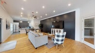 Located at 259 S Maestri Rd in Tontitown, Arkansas, this single-story space presents a thoughtfully designed interior that blends modern farmhouse aesthetics with functional, open-concept living. Natural light hardwood flooring flows throughout the main gathering areas, complementing the crisp white walls and crown molding that frame each room.

The expansive main living area is anchored by a striking floor-to-ceiling black paneled accent wall, which serves as a dramatic backdrop for the entertainment setup. Recessed lighting and a ceiling fan maintain a comfortable, well-lit atmosphere. A secondary seating nook, also set against a matching black panel wall, opens directly to the outdoors through large sliding glass doors, offering unobstructed views of a community pool and surrounding neighborhood.

The kitchen is well-appointed with white shaker cabinetry, gray quartz countertops, stainless steel appliances including a French door refrigerator and dishwasher, and a textured dark hexagon tile backsplash. Large windows flood the kitchen with natural light. Gray tile flooring grounds the space with a clean, contemporary contrast to the bright cabinetry. A dining area and additional lounge seating complete this versatile, open layout.