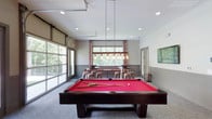 Located at 3130 Hillandale Rd in Durham, North Carolina, this single-floor community amenity space is thoughtfully designed for recreation and social gathering. The space features a contemporary aesthetic with neutral gray tones, wainscoting-style wall paneling, recessed lighting, and plush carpeted floors throughout.

At the heart of the space is a full-sized billiards table with a bold red felt surface, illuminated by an industrial-style pendant light fixture with exposed Edison bulbs. Flanking the billiards area are large windows and a full-length glass garage-style door, flooding the room with natural light and offering views of the surrounding wooded landscape.

The social and dining area features high-top tables paired with copper-toned metal stools with upholstered pink seats, alongside a built-in shuffleboard table positioned beneath large picture windows dressed with patterned curtains. A dedicated lounge nook includes a built-in cushioned bench with accent pillows, a round ottoman, and a wall-mounted flat-screen TV set against a white-painted brick accent wall. Below the TV, sleek white cabinetry with a countertop provides storage and display space, complemented by a mini refrigerator. Vibrant artwork and decorative accessories add pops of color, giving the overall space an energetic yet welcoming atmosphere.