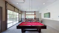 Located at 3130 Hillandale Rd in Durham, North Carolina, this single-floor community amenity space is thoughtfully designed for recreation and social gathering. The space features a contemporary aesthetic with neutral gray tones, wainscoting-style wall paneling, recessed lighting, and plush carpeted floors throughout.

At the heart of the space is a full-sized billiards table with a bold red felt surface, illuminated by an industrial-style pendant light fixture with exposed Edison bulbs. Flanking the billiards area are large windows and a full-length glass garage-style door, flooding the room with natural light and offering views of the surrounding wooded landscape.

The social and dining area features high-top tables paired with copper-toned metal stools with upholstered pink seats, alongside a built-in shuffleboard table positioned beneath large picture windows dressed with patterned curtains. A dedicated lounge nook includes a built-in cushioned bench with accent pillows, a round ottoman, and a wall-mounted flat-screen TV set against a white-painted brick accent wall. Below the TV, sleek white cabinetry with a countertop provides storage and display space, complemented by a mini refrigerator. Vibrant artwork and decorative accessories add pops of color, giving the overall space an energetic yet welcoming atmosphere.