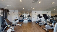 This single-floor fitness center is designed to support a full range of workout routines in a clean, functional environment. The space is outfitted with an impressive lineup of True-brand cardio equipment, including multiple treadmills, upright and recumbent stationary bikes, elliptical trainers, and a multi-station cable strength machine. A set of free weights is also available for supplementary training. The wood-look flooring runs throughout, providing a warm aesthetic contrast to the crisp white walls. Large wall-to-wall mirrors line one side of the room, visually expanding the space and allowing users to monitor their form during workouts. Recessed ceiling lights provide consistent, even illumination throughout the facility. Natural light filters in through windows near the entrance, adding brightness to the room. A dedicated door provides convenient access to the exterior. Fitness center rules are posted on the wall near the entrance, ensuring a well-organized and orderly environment. The overall layout is thoughtfully arranged to maximize usable floor space while keeping equipment accessible and traffic flow unobstructed, making this fitness center a practical and welcoming destination for daily exercise.
