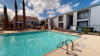 This outdoor amenity space centers around a generously sized swimming pool with clear blue water, marked with depth indicators at the shallow and deeper ends. The pool deck is finished in a light-colored concrete surface and is equipped with metal handrail entry points for easy access. Surrounding the pool, the sun deck is furnished with blue sling-style lounge chairs and umbrella-shaded patio tables and chairs, offering comfortable options for both sunbathing and shaded relaxation. Large terracotta planters serve as decorative accents throughout the deck area, adding a touch of warmth to the overall aesthetic. The space is enclosed by a black wrought-iron fence, providing defined boundaries while maintaining an open, airy feel. Mature trees, including tall cypress and pine varieties, line the perimeter and provide a natural backdrop. The multi-story residential buildings that flank the pool area feature gray lap siding and private balconies, contributing to the communal courtyard atmosphere. A separate patio seating area with additional umbrella tables is visible near a community building, ideal for social gatherings. The entire space benefits from open sky exposure, allowing for abundant natural light throughout the day.