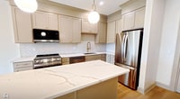 Unit 409 at 983 Michigan Avenue in Buffalo, NY is a thoughtfully designed 600-square-foot, one-bedroom, one-bathroom apartment spanning a single floor. The open-concept kitchen and living area serves as the heart of the unit, showcasing taupe shaker-style cabinetry with gold hardware, a herringbone tile backsplash, quartz countertops, a central island with pendant lighting, and a full suite of stainless steel appliances including a gas range and French door refrigerator. Light hardwood flooring runs throughout the main living areas, complementing the bright, neutral palette. The spacious living room receives natural light through large windows with outdoor views. The 140-square-foot bedroom features plush carpet, a built-in closet with shelving, and a window with natural light. A second flexible room with hardwood floors and a large window offers additional living or workspace potential. The bathroom is distinctively finished with penny-round mosaic tile flooring, bold floor-to-ceiling black tile in the walk-in shower, and black matte hardware accents. A dedicated in-unit laundry closet houses a stacked LG washer and dryer, adding everyday convenience to this well-appointed Buffalo apartment.