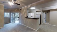 This single-floor space showcases an open-concept design that seamlessly connects the living area and kitchen. The living room is finished with plush wall-to-wall carpeting in a neutral tone, complemented by crisp white crown molding that adds a classic architectural detail. A ceiling fan overhead provides both function and a subtle decorative element, while a sliding glass door floods the space with natural light and opens to a private balcony overlooking mature trees, creating a pleasant indoor-outdoor connection.

The kitchen is defined by a peninsula with a dark countertop, offering a natural divider between the cooking and living spaces while maintaining an open feel. White cabinetry lines the kitchen walls, providing ample storage, and is paired with a full-size refrigerator. The kitchen features tile flooring, contrasting neatly with the carpeted living area. Built-in shelving is also visible, adding functional storage options. A washer and dryer appear to be included within the unit. The overall color palette of warm beige and white tones throughout creates a cohesive, calm, and inviting atmosphere well-suited for everyday living.