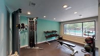 This single-floor fitness center in Marietta, GA, 30064, is thoughtfully designed to accommodate a well-rounded workout experience. The facility is divided into distinct functional zones, each outfitted with specific equipment to support varied training styles. The cardio area features multiple treadmills and an elliptical machine positioned alongside large windows that overlook a landscaped outdoor area, providing natural light and exterior views that enhance the workout environment. A full-length mirror wall in this section visually expands the space and allows for form monitoring during exercise. The strength training zone is painted in a vibrant teal accent color and houses a multi-functional cable machine, a two-tier dumbbell rack with a full range of free weights, a adjustable workout bench, and an assortment of medicine balls. Rubber-tile flooring throughout the facility provides cushioning and durability suitable for high-impact activity. A dedicated open area with foam mat flooring offers space for stretching, yoga, or functional training. A wall-mounted water fountain is conveniently accessible within the facility. Recessed lighting provides consistent illumination throughout, and a window overlooking a fenced pool area connects the indoor and outdoor amenities of the property.