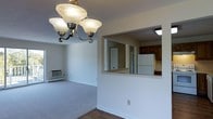 This single-floor space is designed with a practical and straightforward layout. The living area features wall-to-wall carpeting and neutral-toned walls, creating a clean and simple atmosphere. A classic three-bulb chandelier with frosted glass shades serves as a focal point overhead, adding a touch of traditional charm. A wall-mounted heating and air conditioning unit is positioned along one wall, providing climate control directly within the living space.

A defining architectural feature is the open pass-through window connecting the living area to the kitchen. This cutout allows visual connectivity between the two spaces without fully opening the floor plan. The kitchen, visible through the pass-through, includes dark wood cabinetry, a white refrigerator, and additional appliances. A separate door also provides direct access to the kitchen.

The transition from carpeted living space to hard flooring at the kitchen threshold helps define each zone. The overall ambiance is neutral and functional, with ample wall space and a straightforward design that offers flexibility for a variety of furniture arrangements and personal décor styles.