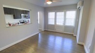 This single-floor residence offers a well-designed, open layout that maximizes both comfort and functionality. The living area is a standout feature, showcasing rich hardwood floors that reflect natural light streaming in through large, white-trimmed windows fitted with blinds. A ceiling fan adds a practical touch overhead, enhancing airflow throughout the space.

The living area flows seamlessly toward a set of glass-paneled double doors that lead to the exterior, filling the space with warmth and an inviting sense of openness. Neutral gray walls complement the light tones throughout, creating a clean and contemporary ambiance.

The kitchen is accessible via a pass-through counter with a granite-style surface, offering a convenient connection between the cooking and living spaces. Dark cabinetry contrasts elegantly with stainless steel appliances, including a microwave and range, providing a modern and efficient cooking environment.

A doorway visible from the main living space suggests additional rooms beyond, contributing to a practical and well-organized floor plan. The combination of natural materials, neutral finishes, and ample natural light makes this a welcoming and versatile space suited to a variety of lifestyles.