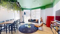 Located at 340 Third St in Jersey City, NJ, this single-floor living space offers a bold and visually dynamic interior design that immediately captures attention. The most striking architectural feature is the vivid green ceiling, which spans the entire space and creates a lively, energetic atmosphere complemented by warm white walls and light hardwood flooring throughout.

A dramatic overhead installation of cascading live plants hangs above a long communal bar-height table with seating for multiple guests, serving as both a functional dining area and a stunning visual centerpiece. Pendant lighting is woven throughout the greenery, providing warm, ambient illumination. Large floor-to-ceiling windows allow generous natural light to flood the space, framing sheer white curtains and offering views to the exterior.

The lounge area features a comfortable gray sofa with accent pillows, a leather chair, and a celestial-themed circular area rug anchoring colorful, mushroom-shaped side tables in lime green and aqua. A bookshelf stocked with an eclectic collection of books adds an intellectual touch near an arched hallway entrance. A retro red SMEG refrigerator and a large flat-screen TV round out the space, alongside decorative cactus accents. Abstract wall art adds a finishing creative flourish throughout.