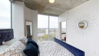 This contemporary high-rise unit showcases a striking industrial-modern aesthetic defined by its exposed concrete ceiling, which adds raw texture and architectural character to the space. The bedroom is a standout feature, dominated by expansive floor-to-ceiling corner windows that flood the room with natural light and frame breathtaking panoramic views of the surrounding cityscape and beyond. The elevated position of the unit offers a wide, unobstructed perspective of the urban landscape, making the views a true focal point of the living experience. White walls provide a clean contrast to the raw concrete overhead, creating a balanced interplay between industrial and modern design elements. The bedroom comfortably accommodates a full-sized bed dressed in patterned bedding accented with rich teal throw pillows, complemented by a decorative starburst wall accent that adds a touch of personality. A full-length mirror enhances the sense of space and reflects the abundant natural light. An additional side window further amplifies the brightness and connection to the outdoors. The overall ambiance is one of urban sophistication, with the interplay of natural light, elevated views, and industrial-modern finishes creating a distinctive and memorable living space.