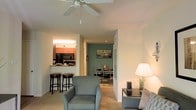 This single-floor residence is designed with an open-concept layout that seamlessly connects the primary living spaces. The living area serves as the central hub, featuring neutral-toned carpeting, soft walls, and a ceiling fan that promotes air circulation and comfort. A plush armchair and sofa furnish the space, creating a relaxed and inviting atmosphere.

Adjacent to the living area, the kitchen is positioned behind a raised breakfast bar with dark-stained bar stools, offering a casual dining option. The kitchen features warm wood cabinetry, under-cabinet lighting, and dark appliances, including a built-in microwave, lending a practical and efficient workspace.

A doorway from the living area leads into the dining room, which is distinguished by a muted sage-green accent wall that adds a subtle contrast to the otherwise neutral palette. A dining table with seating is centrally placed in this room, making it well-suited for meals and gatherings.

The overall ambiance of the space is calm and functional, with consistent flooring and a cohesive color scheme tying the rooms together. The layout supports a smooth flow between living, cooking, and dining areas.