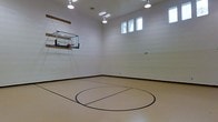 This single-story indoor basketball court offers a dedicated athletic space designed for recreational play. The court features a regulation-style wall-mounted glass backboard and hoop, positioned on one end wall, with clearly painted court markings — including a prominent center circle — laid over a smooth, light-toned sport flooring surface. The perimeter walls are finished in clean, neutral tones with a dark baseboard accent that visually grounds the space. Overhead, a series of recessed and track-mounted lights provide consistent, even illumination across the entire playing surface. A row of clerestory windows along one upper wall introduces natural light, enhancing the brightness of the interior while maintaining a focused athletic environment. The soaring ceiling height accommodates full-range play without obstruction, and the uncluttered wall surfaces contribute to a clean, functional aesthetic. The overall design prioritizes utility and performance, making this an excellent private sports facility suited for basketball practice, recreational games, or multi-sport use.