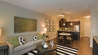 This single-floor residence offers a thoughtfully designed open-concept layout that brings the living, dining, and kitchen spaces together in a cohesive and functional flow. The living area is anchored by a comfortable neutral-toned sofa accented with bold decorative pillows, complemented by a classic round wooden coffee table and a striking black-and-white striped area rug that adds graphic contrast to the warm hardwood floors. A large metallic wall art piece serves as a dramatic focal point, lending a touch of elegance to the space. Adjacent to the living area, the dining space accommodates a round table with wicker-style chairs, creating an intimate setting ideal for everyday meals or entertaining. The kitchen features rich dark cabinetry, a kitchen island with pendant lighting overhead, and stainless steel appliances, all illuminated by stylish track lighting that highlights the space's modern sensibility. Crown molding runs throughout, adding a refined architectural detail to the warm, neutral-toned walls. The seamless transition between areas enhances the overall sense of openness, making this residence feel welcoming, functional, and elegantly appointed from every angle.