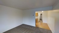 This single-floor space features a straightforward, functional layout designed for comfortable everyday living. The main living area is finished with neutral wall-to-wall carpeting in a warm gray-brown tone, complemented by clean, white-painted walls that create a bright, open feel. A wall-mounted light switch is visible, and the room flows naturally into the adjacent kitchen and hallway areas.

The kitchen, partially visible from the main living space, features warm honey-toned wooden cabinetry and light-colored countertops, with hardwood-style flooring that contrasts with the carpeted living area. The wood tones of the cabinetry lend a classic, traditional aesthetic to the kitchen space.

A hallway extends beyond the kitchen, illuminated by a ceiling-mounted light fixture. The hallway appears to lead to additional rooms, with what seems to be a closet or storage space visible along its length.

Overall, the space maintains a clean, neutral palette throughout, with consistent white walls and simple finishes that provide a versatile backdrop. The single-floor layout offers convenient, accessible living with a practical flow between the main areas.