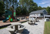 Located in Sandy Springs, GA, this single-story amenity facility offers residents a comprehensive fitness and outdoor recreation experience. The fitness center is thoughtfully designed with two interconnected workout areas, distinguished by bright teal accent walls contrasted against clean white finishes. Rubber flooring throughout provides a safe, durable surface suited for a full range of exercise activities. The space is outfitted with Matrix cardio equipment, including treadmills, ellipticals, and a recumbent bike, alongside a multi-station cable machine, strength training equipment, and a full rack of dumbbells. A wall-mounted TV keeps residents entertained during workouts, while a water station adds convenience. Multiple windows allow ample natural light to fill the space, and French doors open directly to the outdoors. The exterior patio area extends the amenity experience with a paved courtyard featuring modern white lounge furniture, bistro tables, and bar-height seating positioned near a built-in outdoor grill and prep station. Surrounded by mature trees and lush greenery, the outdoor space provides a serene setting for relaxation and socializing, with an elevated deck visible nearby adding architectural interest to the wooded backdrop.