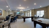 This single-floor communal space is thoughtfully designed to offer a versatile and welcoming environment for relaxation and recreation. The open-concept layout is anchored by light wood-style flooring that runs throughout, lending the space a clean, contemporary feel. Two full-sized billiards tables are prominently positioned at the center of the room, serving as the focal point of the recreational area. A comfortable lounge section features a neutral-toned sofa and seating arrangement situated beneath an elegant chandelier, complemented by a large wall-mounted flat-screen TV and a display shelving unit. Adjacent to the lounge, a dedicated dining area offers a dark-wood table paired with navy upholstered chairs, ideal for gathering or casual meetings. Recessed lighting throughout the ceiling, in addition to the chandelier, ensures bright and even illumination. Large windows along the perimeter walls flood the interior with abundant natural light while providing views to the exterior. Artwork and decorative accents are thoughtfully placed throughout, adding visual interest and a polished finish to the overall ambiance.
