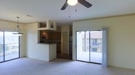 This single-floor residence features an open-concept design that seamlessly connects the living area and kitchen. The carpeted living room is anchored by a ceiling fan with a light fixture overhead, contributing to a comfortable and functional atmosphere. A sliding glass door floods the space with natural light and provides direct access to a private balcony, offering views of the surrounding building exterior.

The kitchen is positioned adjacent to the living area and is partially separated by a raised breakfast bar with a dark stone-style countertop, creating a natural divide while maintaining an open feel. Cabinetry in a warm wood tone lines the kitchen walls, complemented by a stainless steel refrigerator and a built-in microwave. An overhead cabinet structure frames the kitchen entry, adding architectural definition to the space.

A door visible within the kitchen area suggests access to additional rooms or storage. The overall color palette is neutral, with light-toned walls and flooring that create a clean, bright interior. The layout is practical and well-organized, making efficient use of the available space throughout.