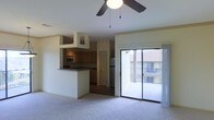 This single-floor residence features an open-concept design that seamlessly connects the living area and kitchen. The carpeted living room is anchored by a ceiling fan with a light fixture overhead, contributing to a comfortable and functional atmosphere. A sliding glass door floods the space with natural light and provides direct access to a private balcony, offering views of the surrounding building exterior.

The kitchen is positioned adjacent to the living area and is partially separated by a raised breakfast bar with a dark stone-style countertop, creating a natural divide while maintaining an open feel. Cabinetry in a warm wood tone lines the kitchen walls, complemented by a stainless steel refrigerator and a built-in microwave. An overhead cabinet structure frames the kitchen entry, adding architectural definition to the space.

A door visible within the kitchen area suggests access to additional rooms or storage. The overall color palette is neutral, with light-toned walls and flooring that create a clean, bright interior. The layout is practical and well-organized, making efficient use of the available space throughout.